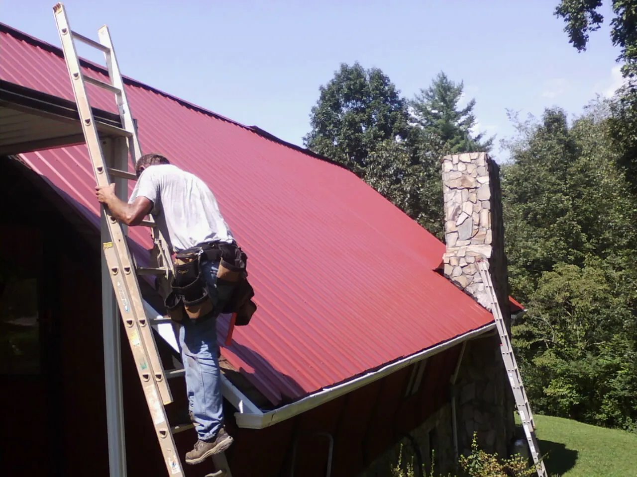 Roofer on a ladder beside a red metal roof and stone chimney — Soffit & Fascia Repair by Roofing Brothers in Oak Creek